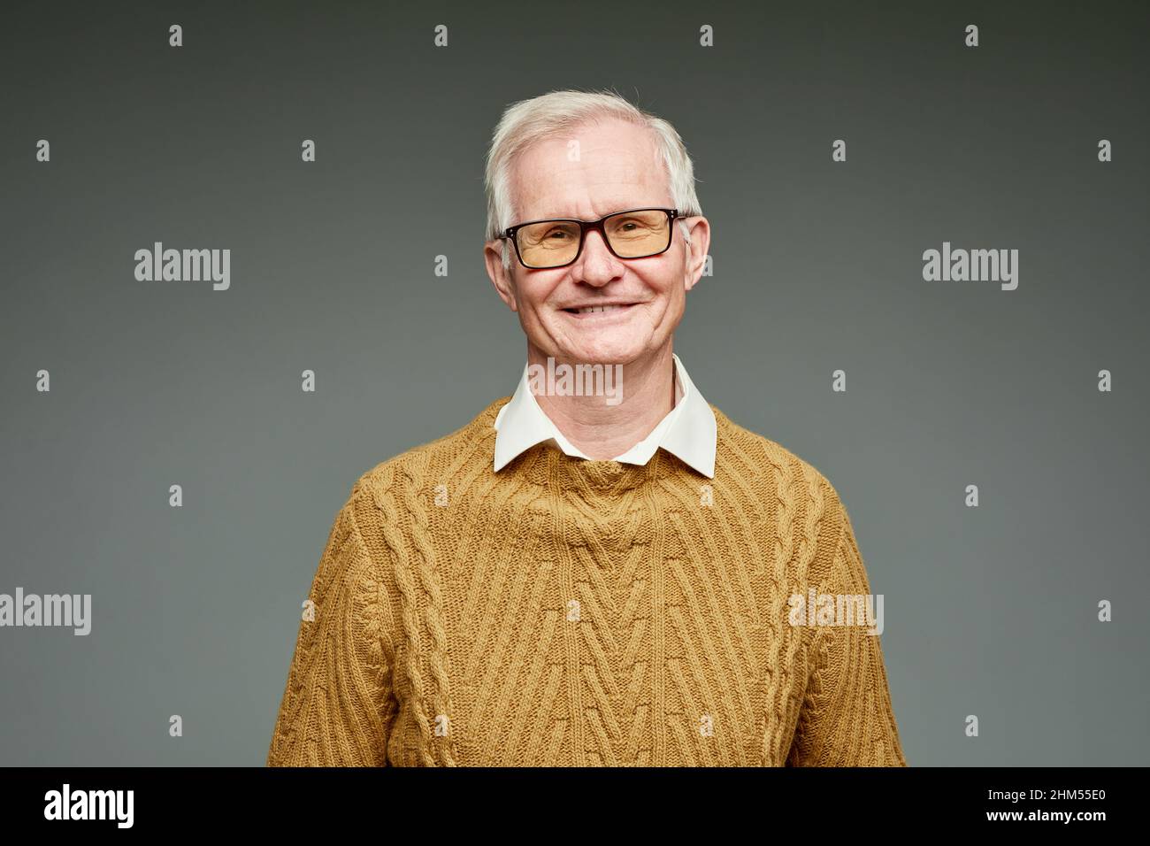 Fröhlicher grauhaariger Mann im Alter mit braunem Strickpullover und Brillen, der vor grauem Hintergrund steht Stockfoto