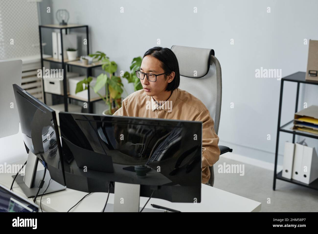 Junger asiatischer Geschäftsmann in Brillen und legerer Kleidung, der am Computermonitor in einem modernen Büro mit offenem Raum sitzt Stockfoto