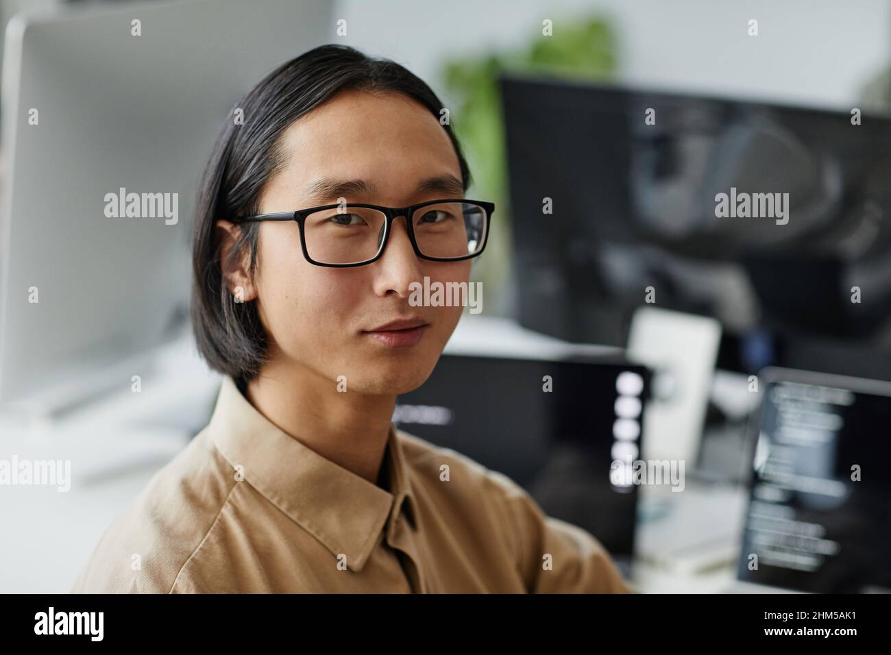 Junger asiatischer Geschäftsmann mit Brille und formellem Hemd, der in einem modernen Büro am Arbeitsplatz auf die Kamera schaut Stockfoto