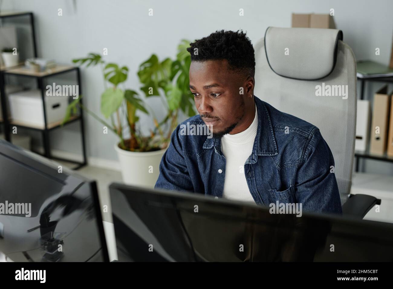 Der junge blackman sitzt in legerer Kleidung vor Computermonitoren und schaut sich während der Arbeit im Büro im Freiraum die Daten auf Bildschirmen an Stockfoto