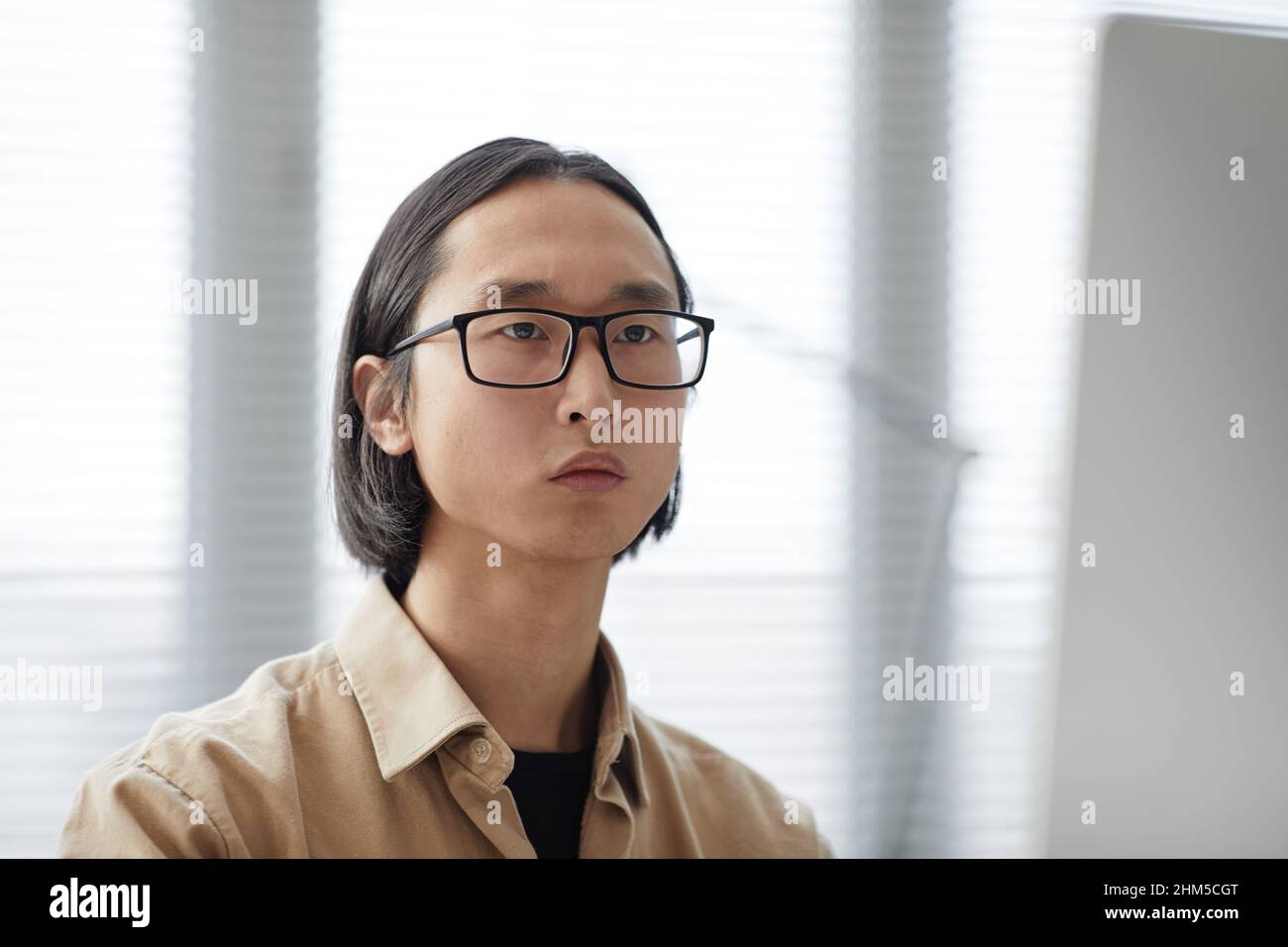 Junge, zeitgenössische männliche Angestellte mit weißem Kragen und asiatischer Ethnie in Brillen und legerem Hemd, die während der Arbeit nach vorne schauen Stockfoto