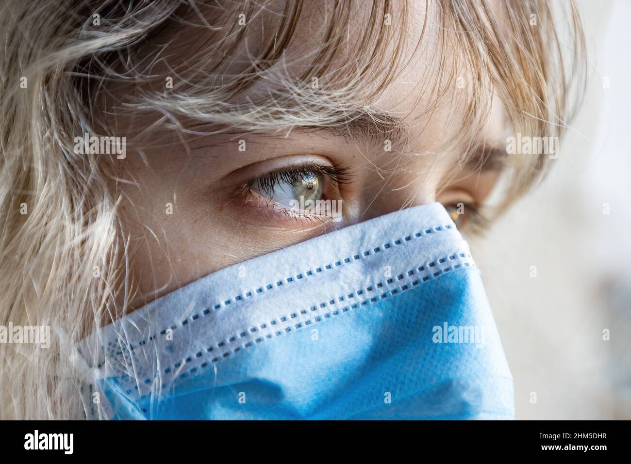 Eine junge Frau, die durch das Coronavirus Covid-19 isoliert wurde, schaut mit einer schützenden Gesichtsmaske durch das Glas ihres Hauses Stockfoto