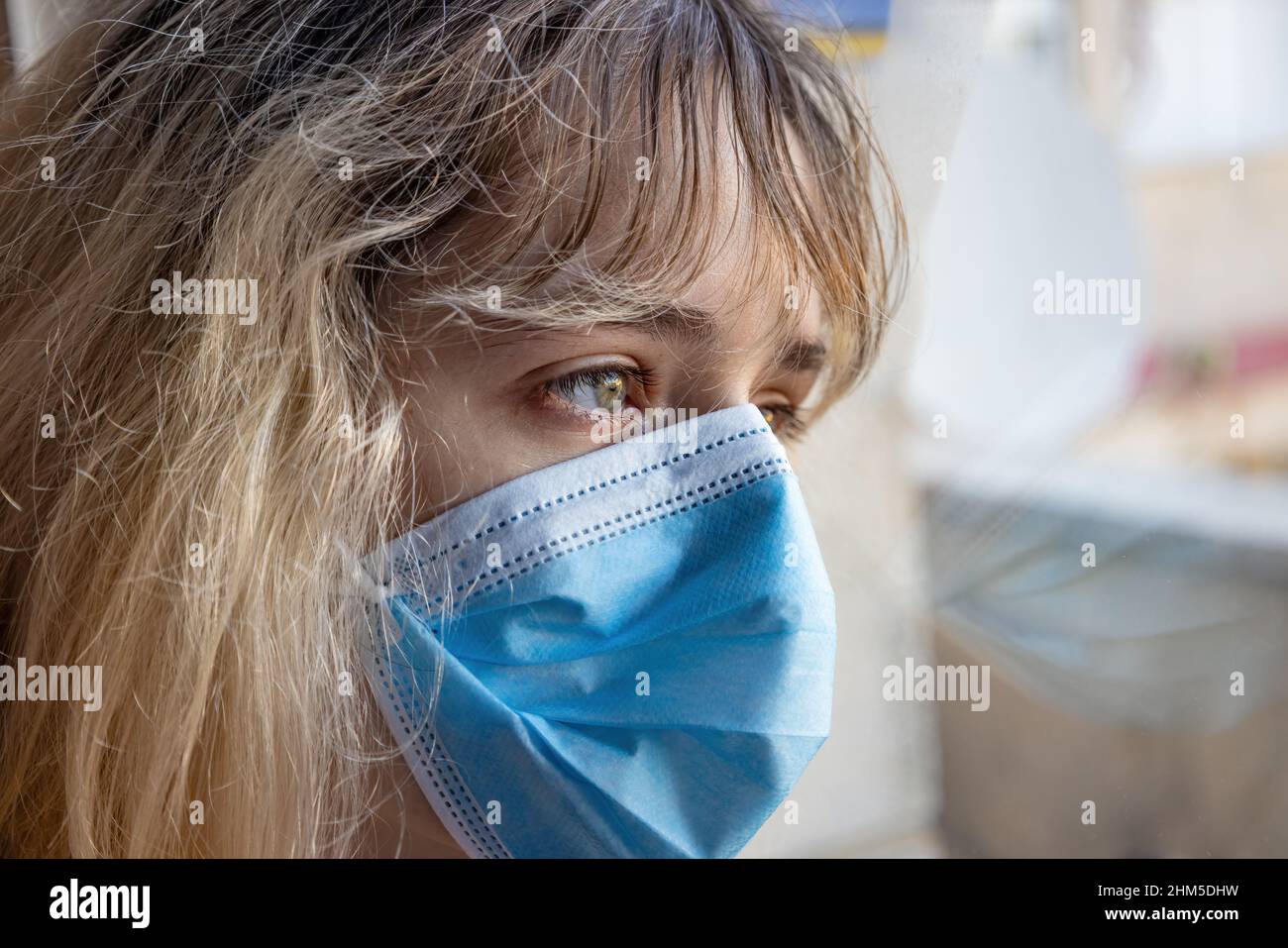Eine junge Frau, die durch das Coronavirus Covid-19 isoliert wurde, schaut mit einer schützenden Gesichtsmaske durch das Glas ihres Hauses Stockfoto