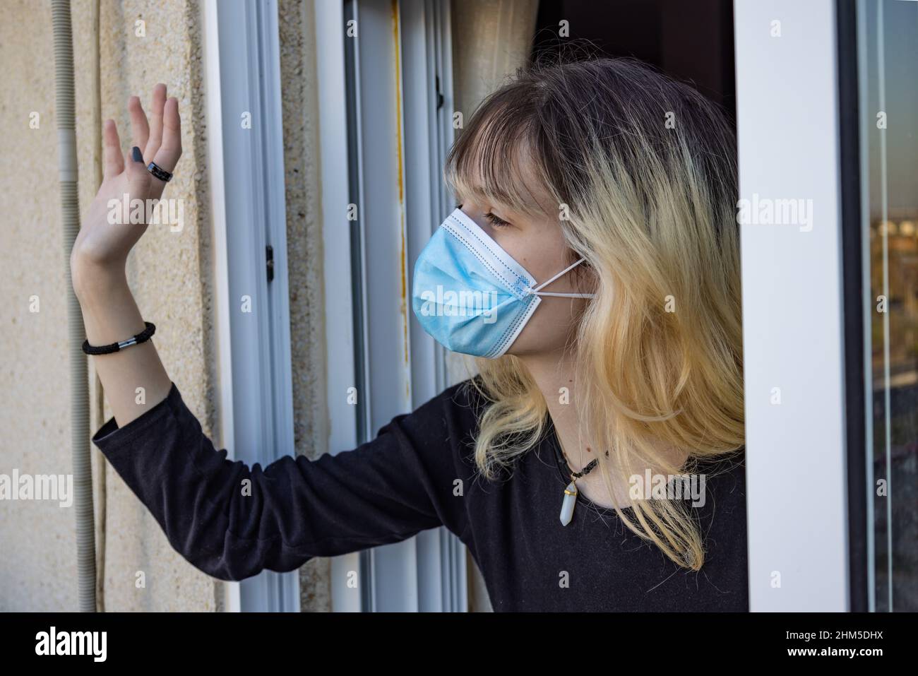 Eine junge Frau, die durch das Coronavirus Covid-19 isoliert wurde, winkt durch ihr Hausfenster und trägt eine schützende Gesichtsmaske Stockfoto