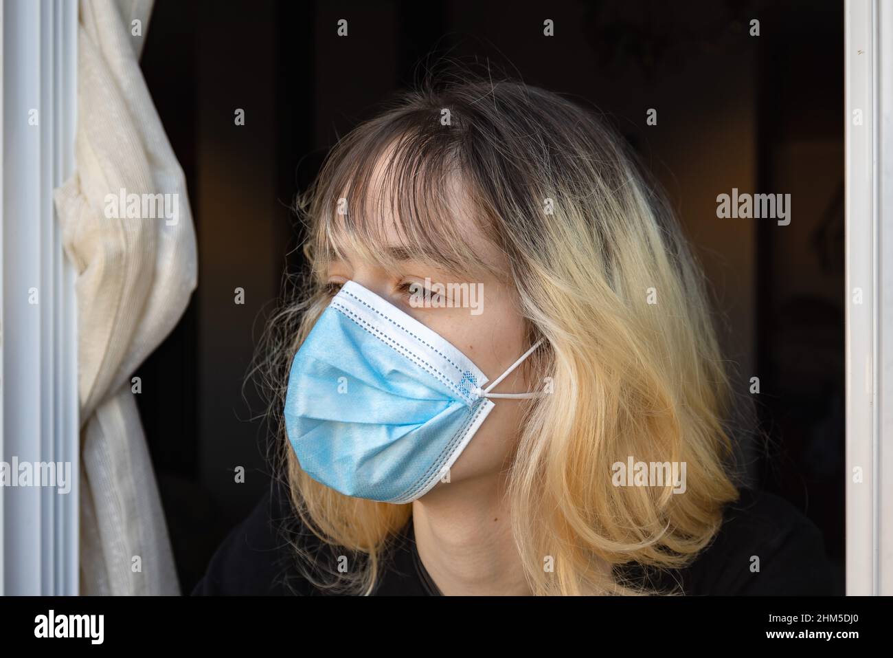 Eine junge Frau, die durch das Coronavirus Covid-19 isoliert wurde, blickt mit einer schützenden Gesichtsmaske durch das Fenster ihres Hauses Stockfoto