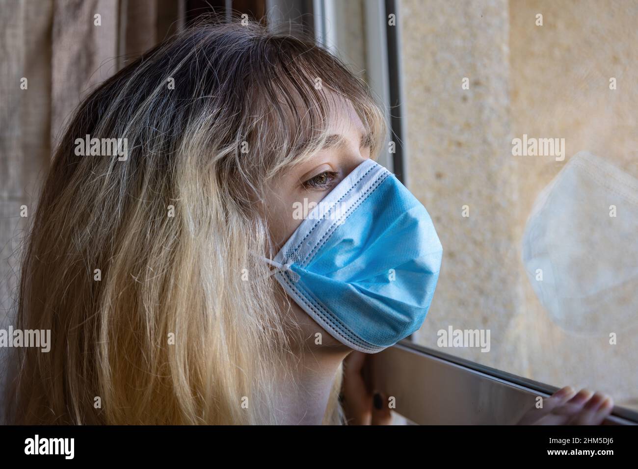 Eine junge Frau, die durch das Coronavirus Covid-19 isoliert wurde, schaut mit einer schützenden Gesichtsmaske durch das Glas ihres Hauses Stockfoto