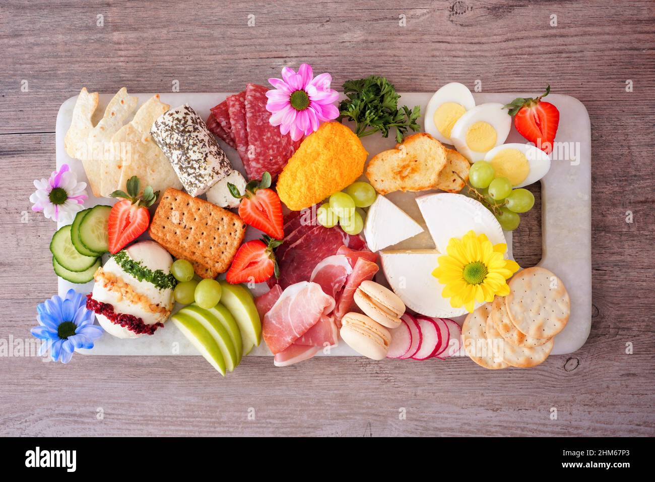 Charcuterie-Tafel mit Frühlings- oder Ostermotiven vor einem Holzhintergrund. Verschiedene Vorspeisen mit Käse, Fleisch, Obst und Gemüse. Ansicht von oben. Stockfoto