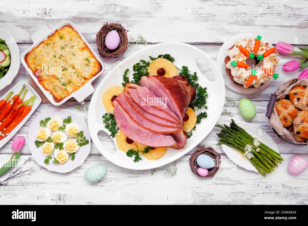 Klassisches Osterschinken-Abendessen. Draufsicht Tischszene auf weißem Holzhintergrund. Schinken, Jakobskartoffeln, Eier, heiße Brötchen, Karottenkuchen und Vegeta Stockfoto