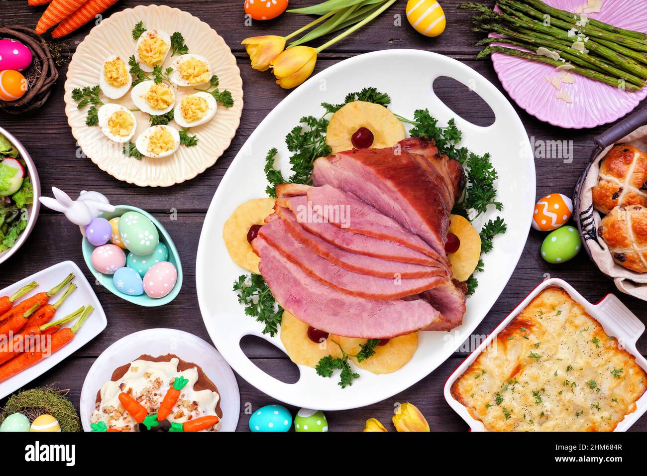 Traditionelles Abendessen mit Osterschinken. Oben Tischszene auf dunklem Holzhintergrund. Schinken, Jakobskartoffeln, Eier, heiße Brötchen, Karottenkuchen und Vegeta Stockfoto