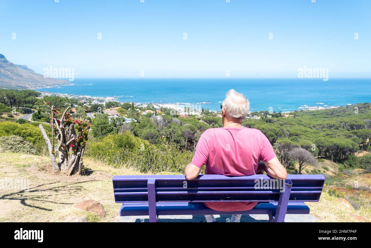 Ein älterer, älterer oder alter kaukasischer Mann sitzt auf einer Holzbank und genießt von einem Hügel in Camps Bay, Kapstadt, die malerische Aussicht auf die Küste Stockfoto