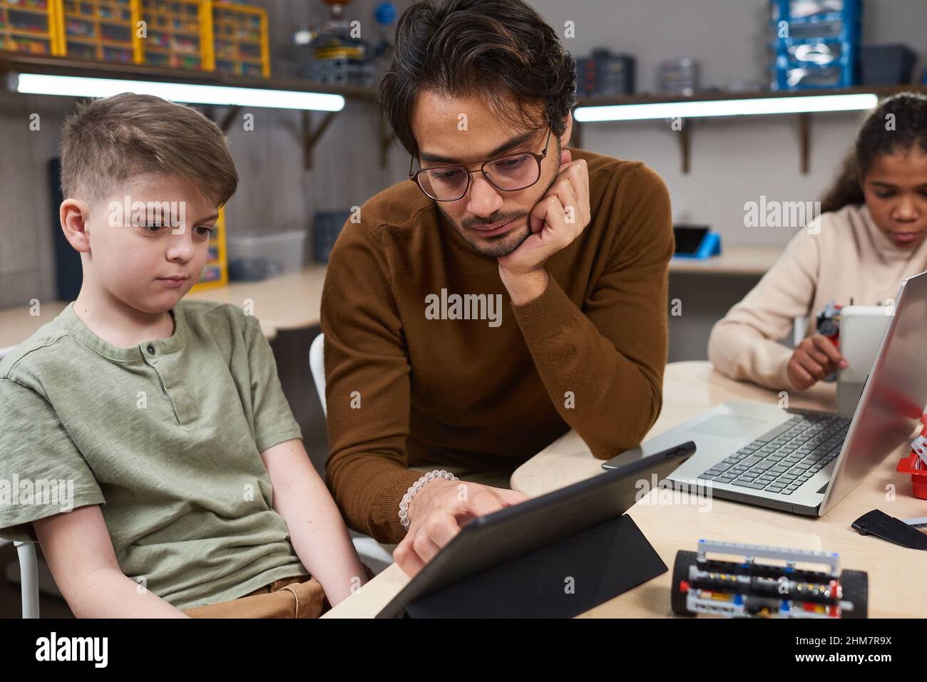 Porträt eines jungen Mannes, der ein digitales Tablet verwendet, während er in der modernen Schule verschiedenen Kindergruppen Roboterunterricht unterrichtete Stockfoto