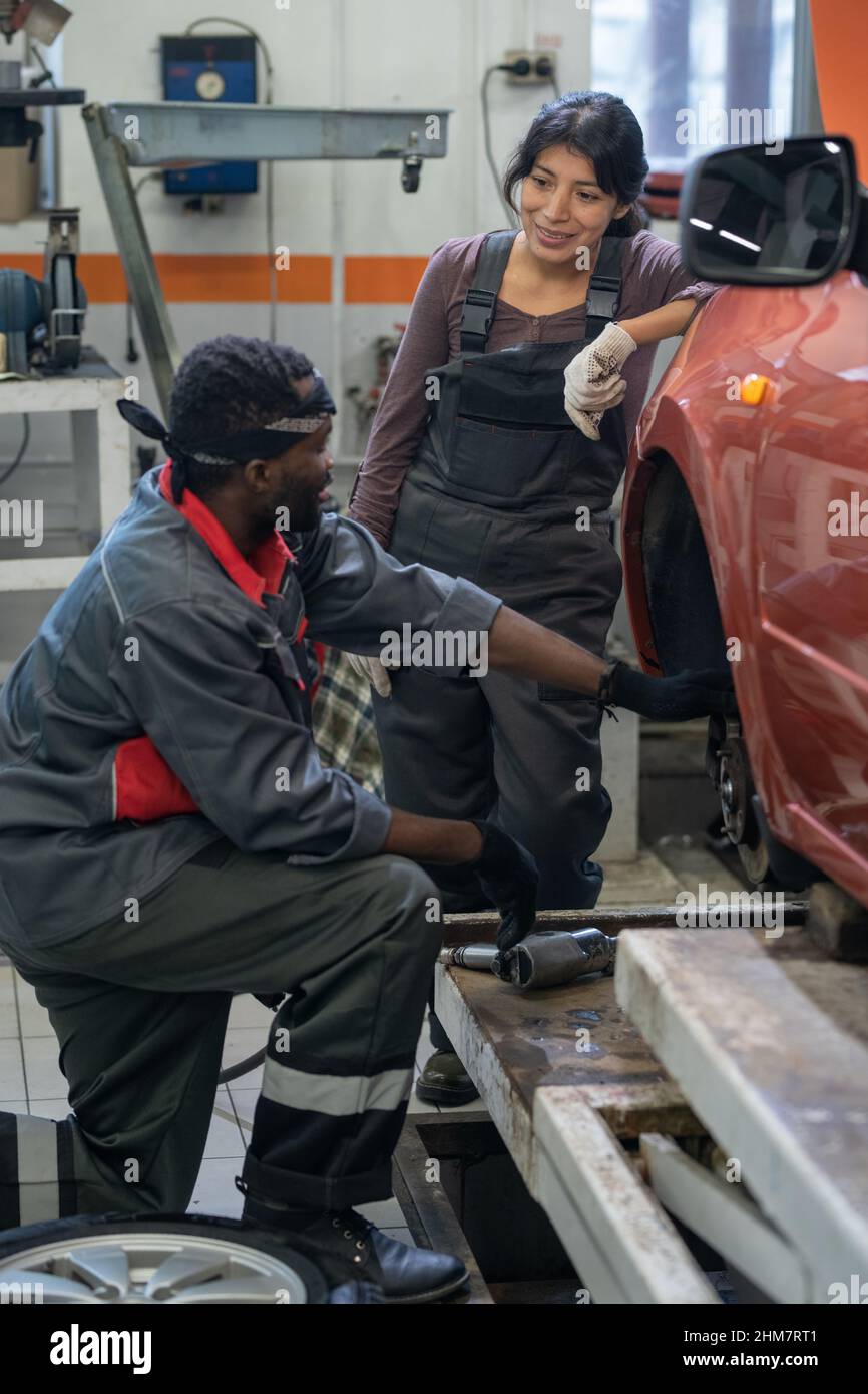 Vertikales Porträt von zwei ethnischen Mechanikern, die das Auto in einer Garage reparieren, und Fokus auf lächelnde junge Frau, die Arbeitskleidung trägt Stockfoto