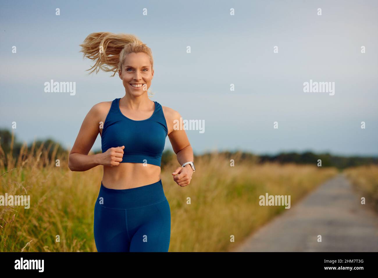 Fit gesunde junge Frau genießen ein Joggen entlang einer Landstraße vorbei an der Kamera mit einem glücklichen Lächeln voller Vitalität in einem aktiven Lifestyle-Konzept Stockfoto