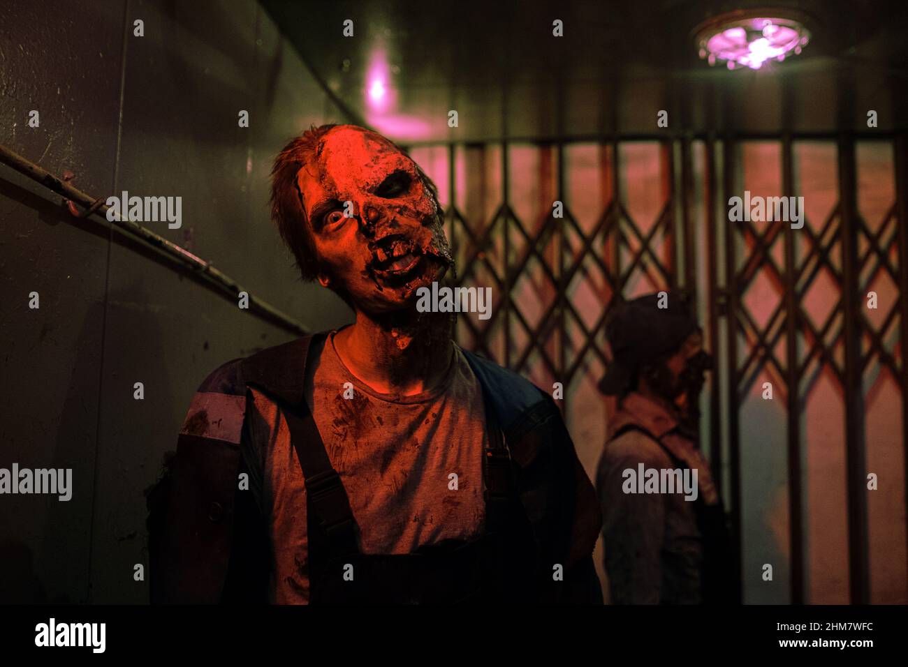 Aufnahme eines männlichen Zombies mit gruseligen Gesichtern, der in einem dunklen, von rotem Licht beleuchteten Flur zur Kamera läuft, um Platz zu kopieren Stockfoto