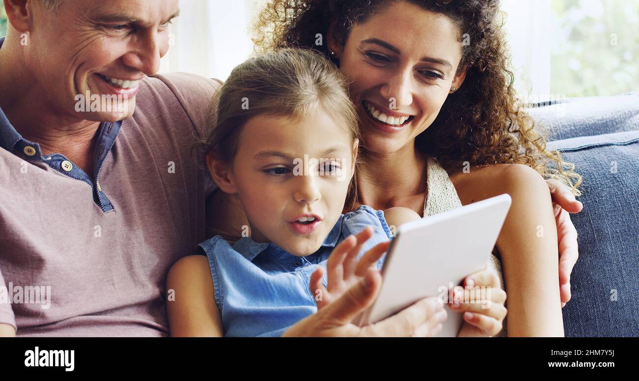 Die Zeit mit der Familie zu einem interaktiven Erlebnis machen. Aufnahme eines niedlichen kleinen Mädchens, das mit ihrer Mutter und ihrem Vater ein digitales Tablet auf dem Sofa zu Hause benutzt. Stockfoto