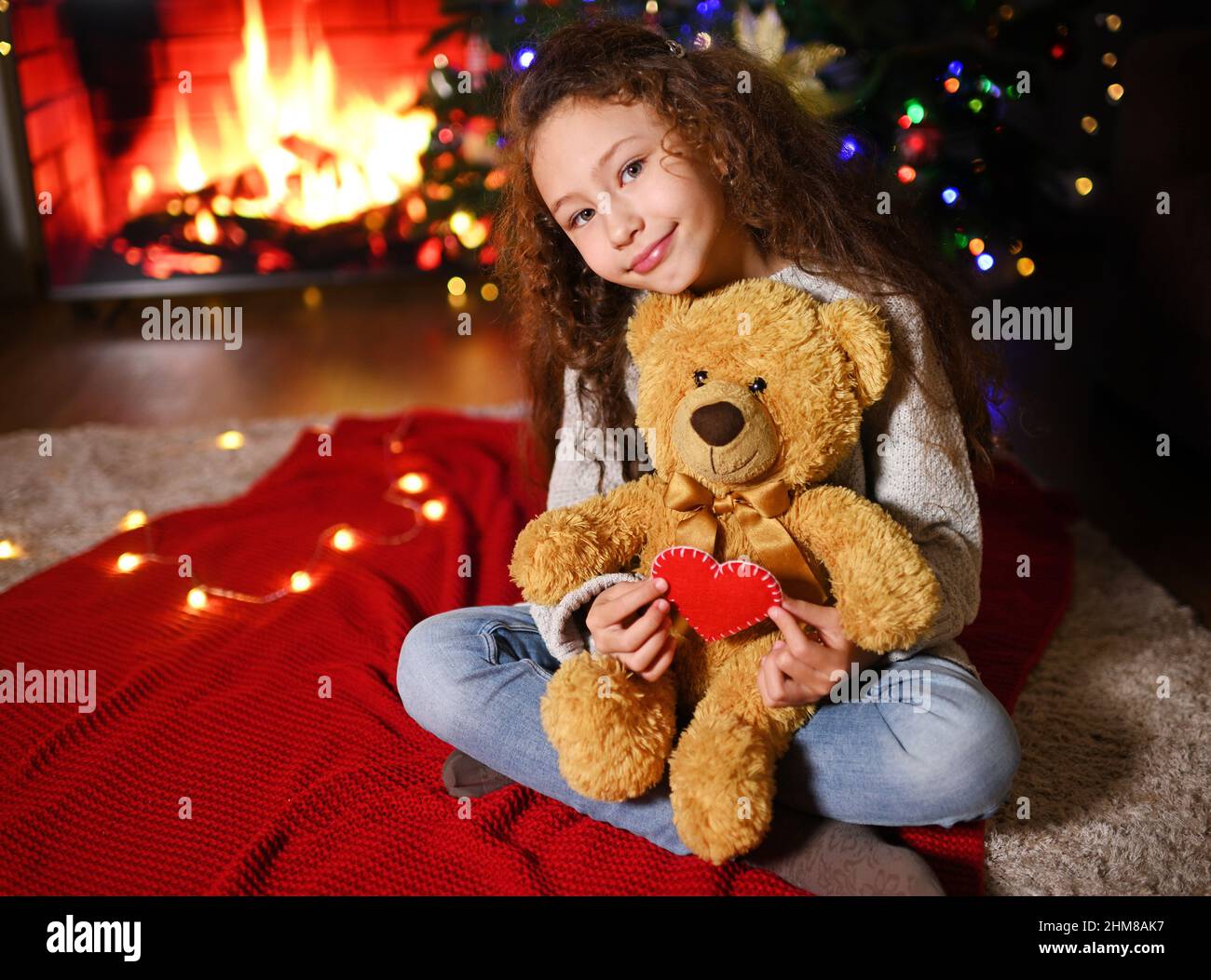 Das Kind ist ein süßes Mädchen mit lockigen Haaren, die lächeln und auf einem roten Karli sitzen und einen Teddybär und ein rotes Herz vor dem Hintergrund der halten Stockfoto
