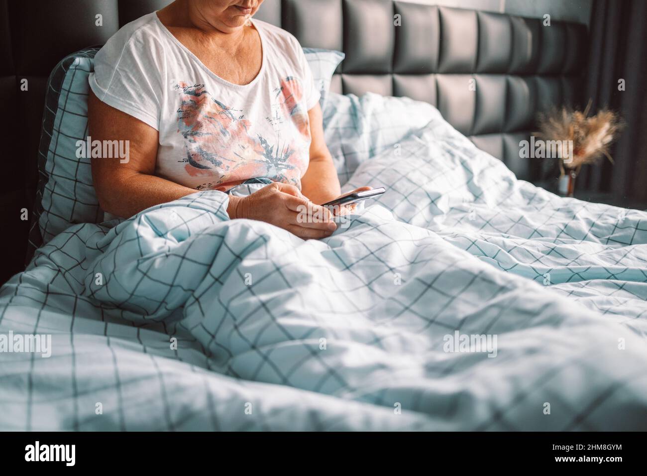 Die blonde Frau mittleren Alters benutzt das Telefon, das im Schlafzimmer auf dem Bett sitzt. Konzept „Guten Morgen, wach auf“ Stockfoto