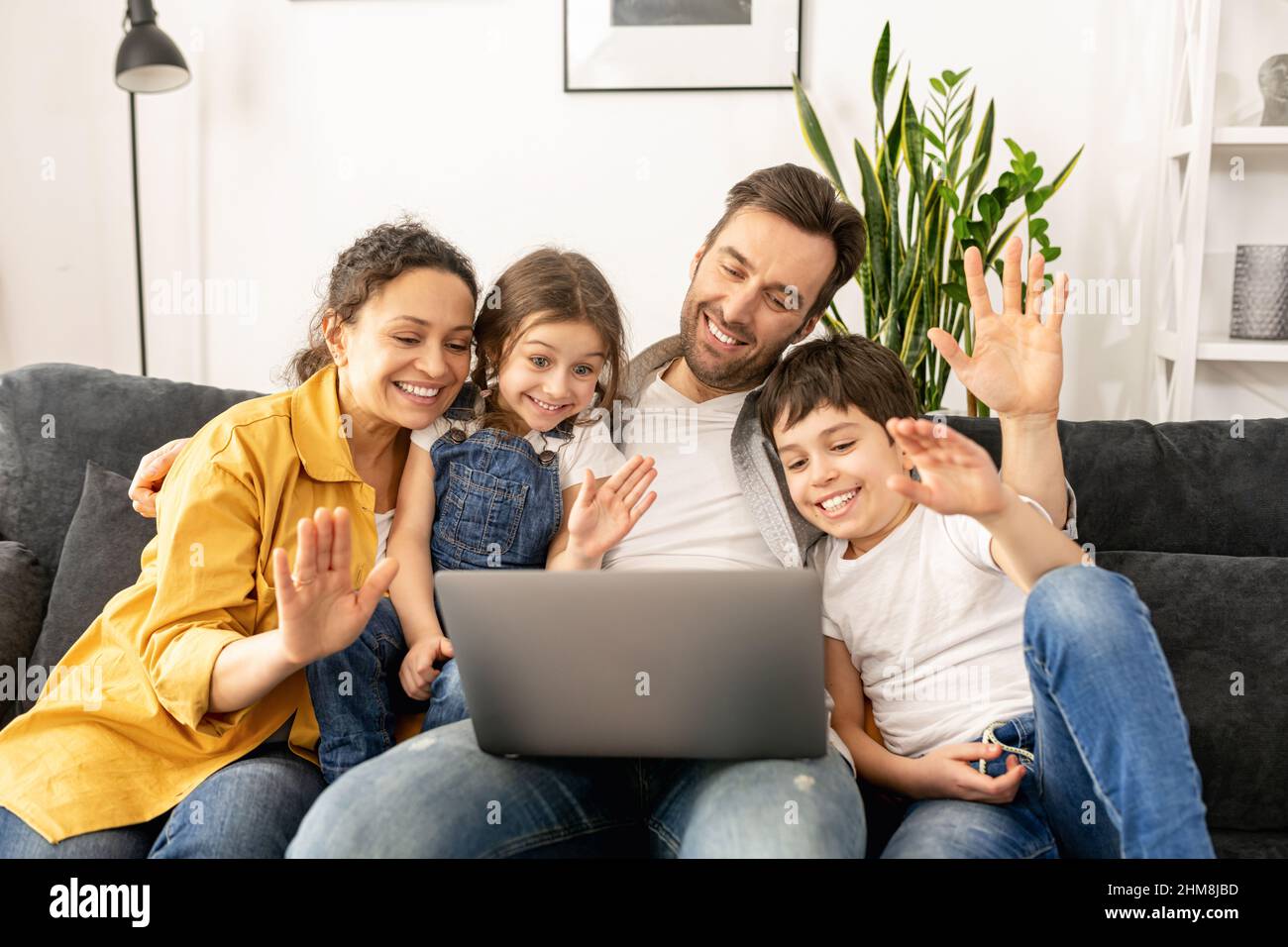Vierköpfige Familie mit Laptop für Videoverbindung mit Großeltern oder Familie. Multirassische Mutter, Vater und zwei Kinder winken in der Webcam des Laptops, Videoanruf, virtuelles Konferenzkonzept Stockfoto