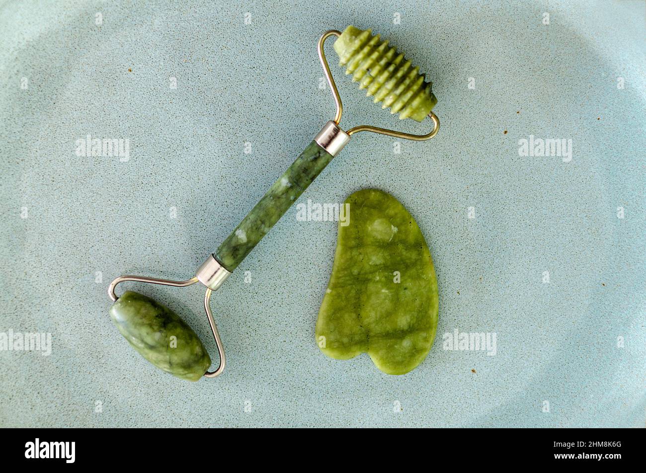 Green Face Roller und Gua Sha Massagegerät. Jade-Walze auf blauem Hintergrund isoliert. Konzept der Gesichtsmassage. Stockfoto