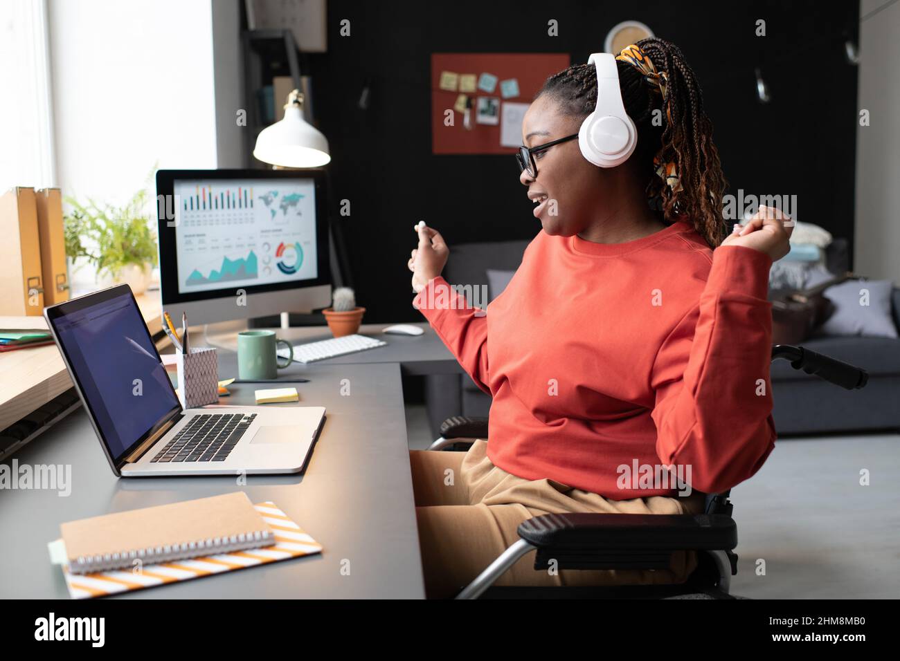 Afrikanische Frau, die während ihrer Arbeit zu Hause am Tisch vor dem Laptop sitzt und Musik hört und tanzt Stockfoto