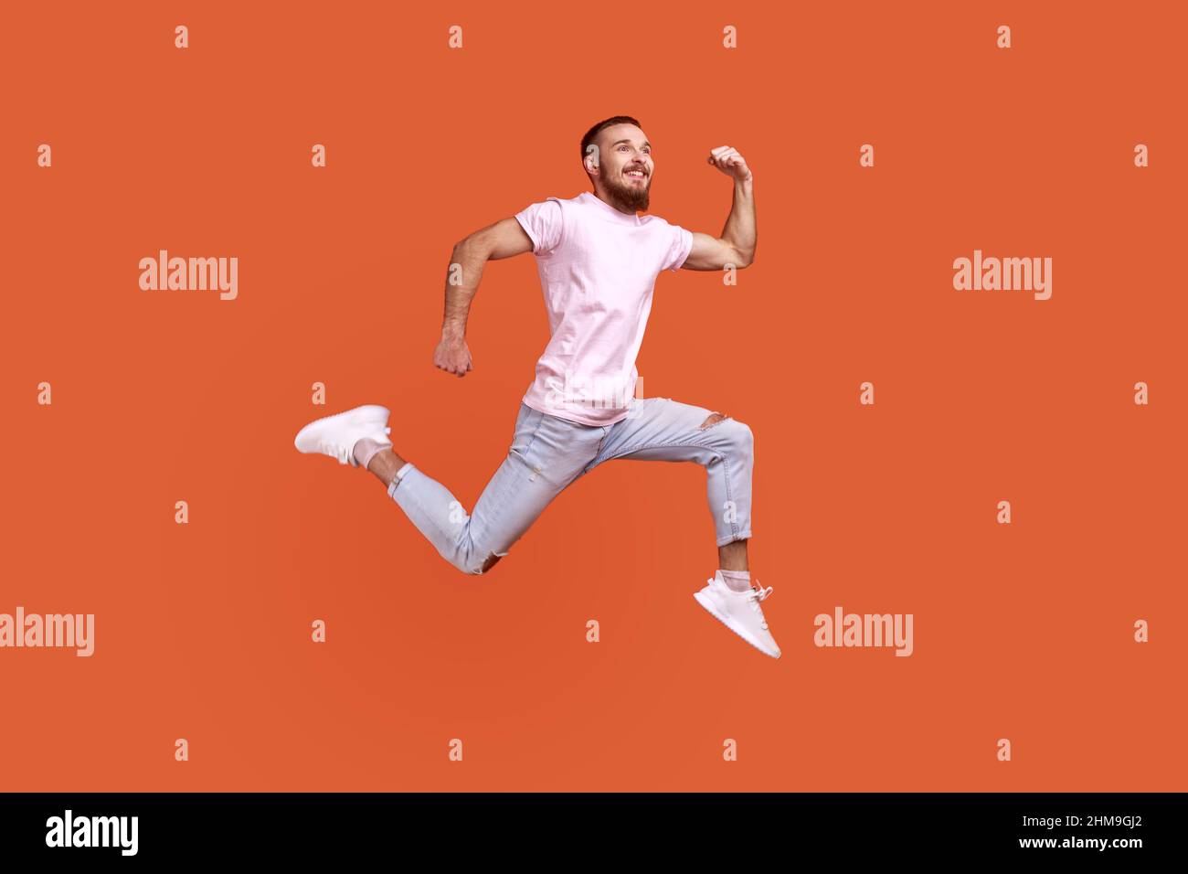 Ganzkörperportrait mit positiv inspiriertem Bartmann, der in der Luft springt oder schnell läuft und Glück zum Ausdruck bringt, in einem rosa T-Shirt. Innenaufnahme des Studios isoliert auf orangefarbenem Hintergrund. Stockfoto