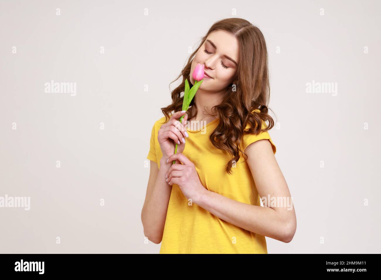 Porträt eines glücklichen verträumten Teenager-Mädchens in gelbem T-Shirt riechende rosa Tulpe mit geschlossenem Auge, lächelnd vor Freude, Frühlingsstimmung, 8. März Frauentag. Innenaufnahme des Studios isoliert auf grauem Hintergrund. Stockfoto