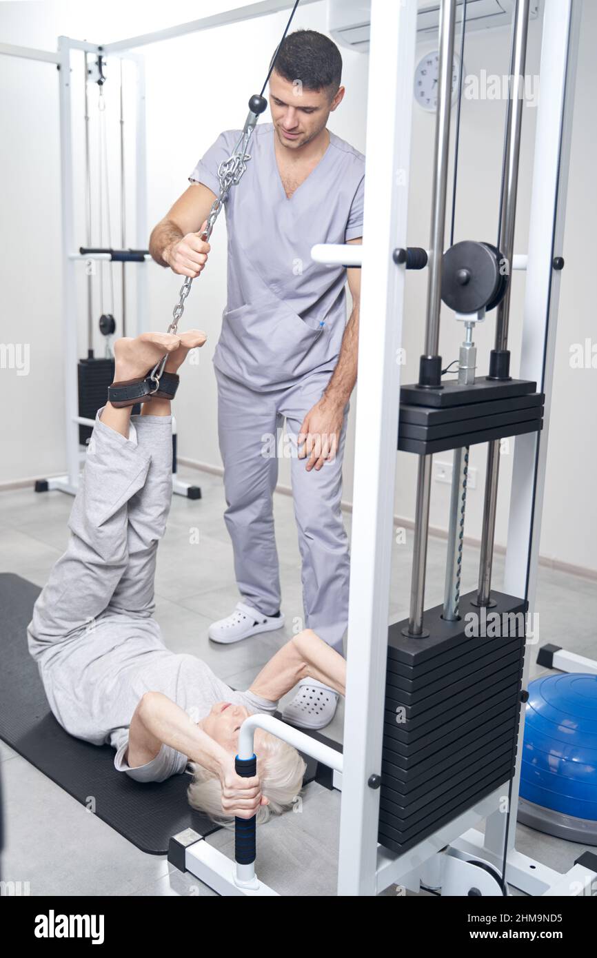 Ältere Frau, die unter Aufsicht eines Physiotherapeuten die Beine mit einem Kabelgerät hochhebt Stockfoto
