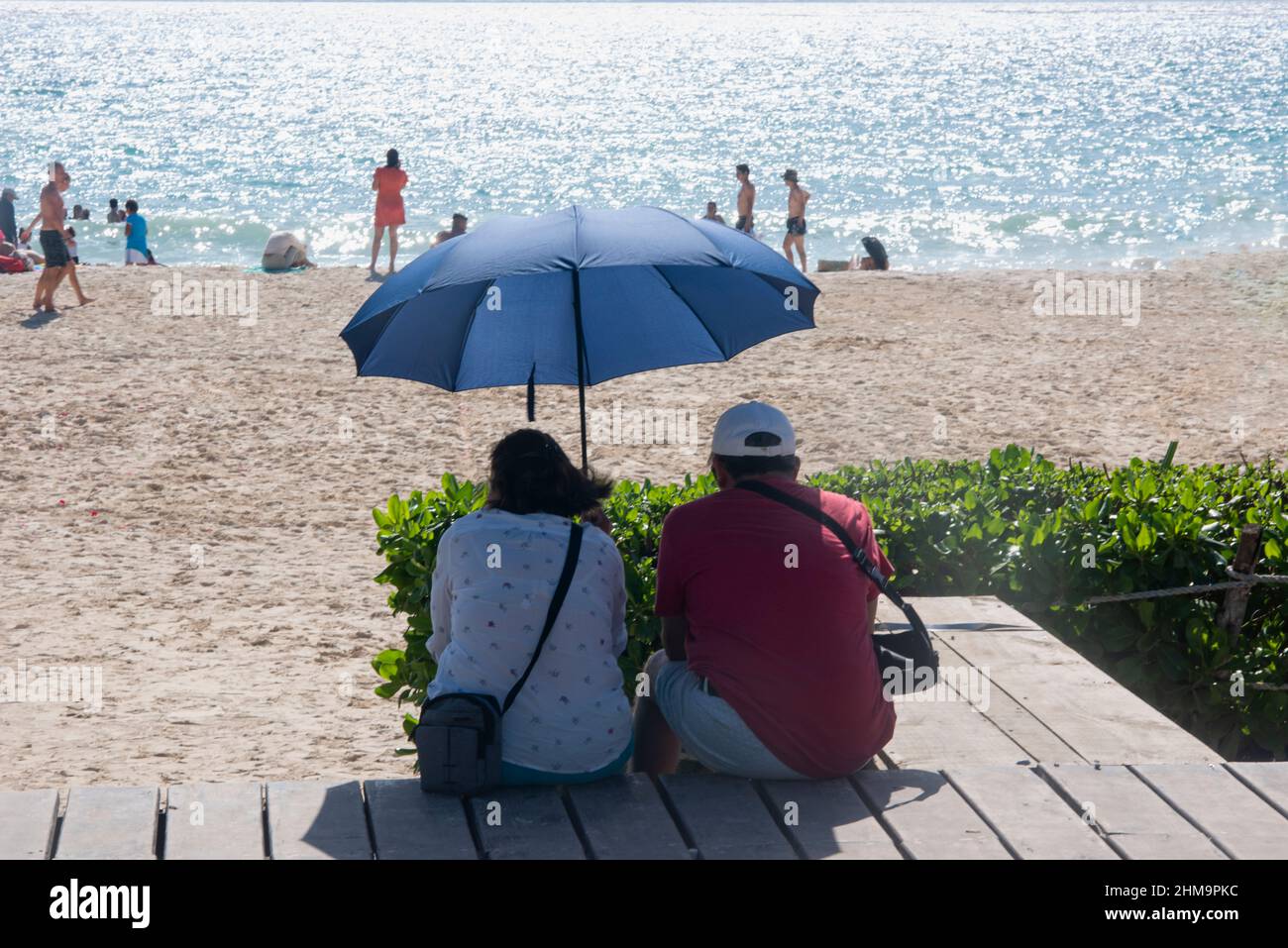 Ein Latino-Touristenpaar beobachtet das Karibische Meer vom Strand aus, während es sich in Playa del Carmen, Mexiko, mit einem Sonnenschirm vor den Sonnenstrahlen schützt. Stockfoto