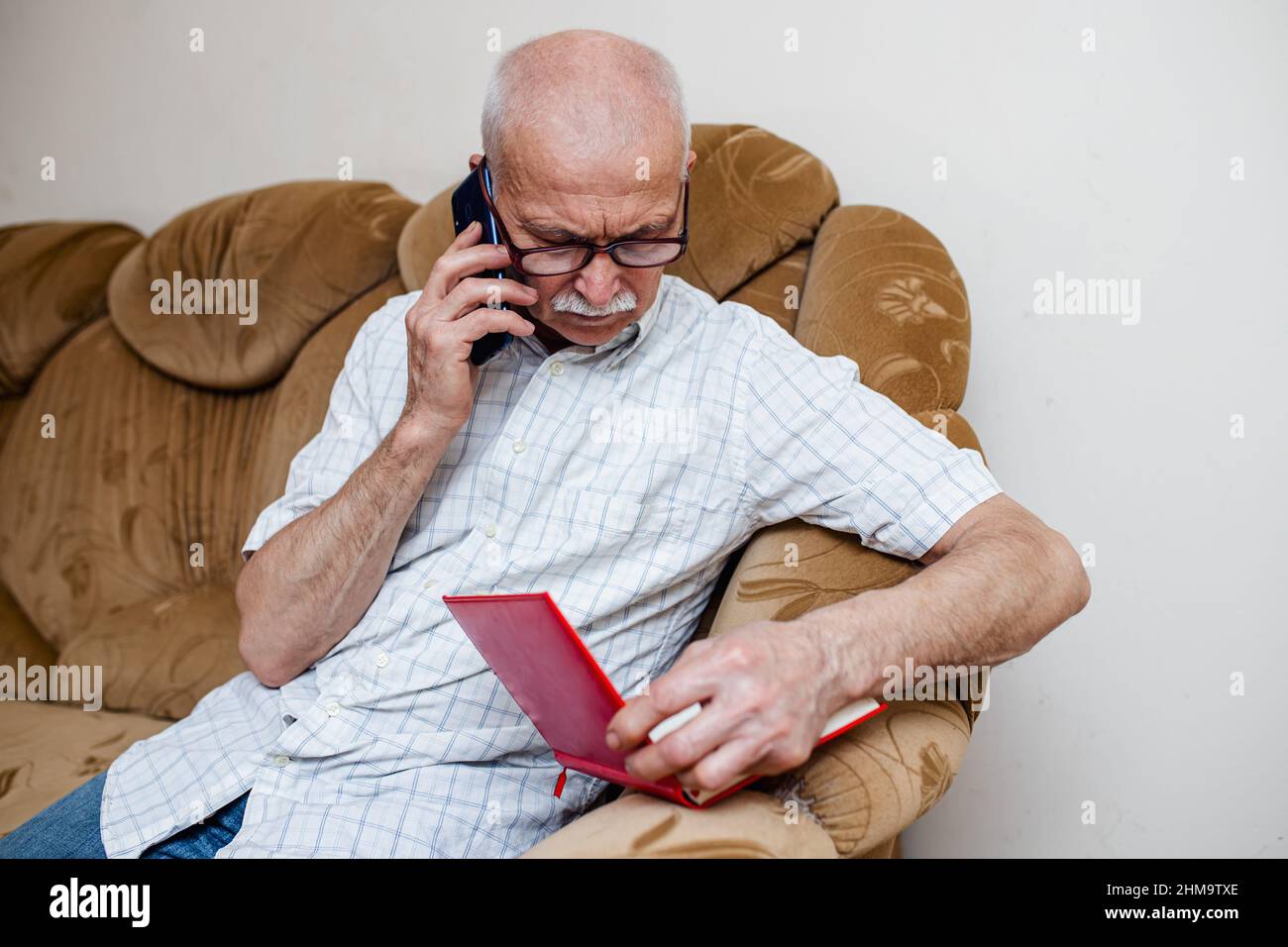 Ein älterer Mann spricht auf seinem Handy und überprüft Daten in einem Notizbuch. Ein alter Geschäftsmann plant Einkäufe und ruft im Geschäft an. Senioren planen Ausgaben Stockfoto