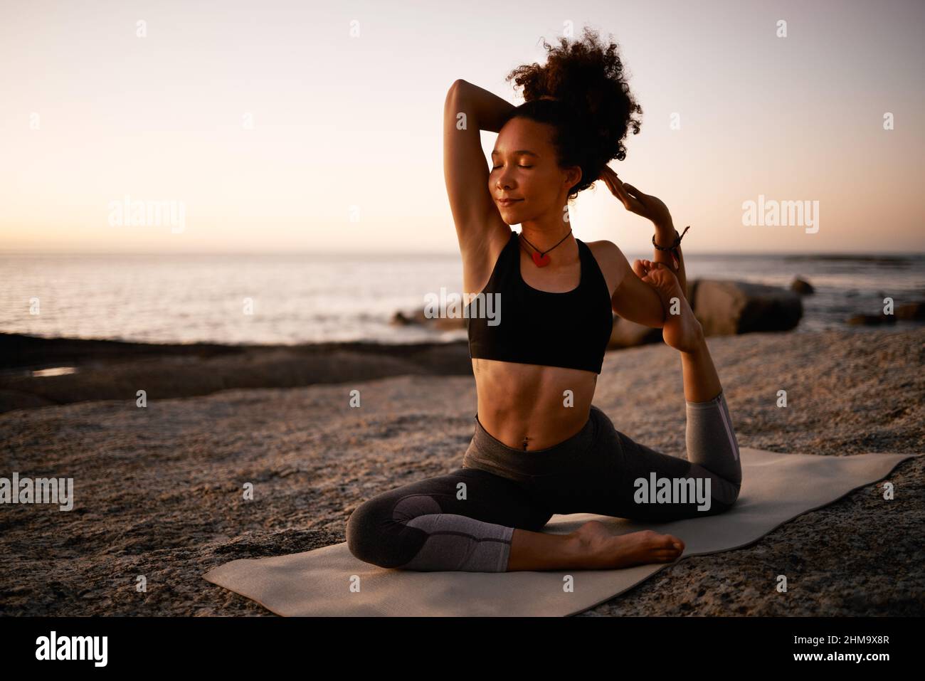 Yoga. Gut für den Geist, gut für den Körper. Ganzkörperaufnahme einer attraktiven jungen Frau, die am Strand bei Sonnenuntergang Yoga praktiziert. Stockfoto