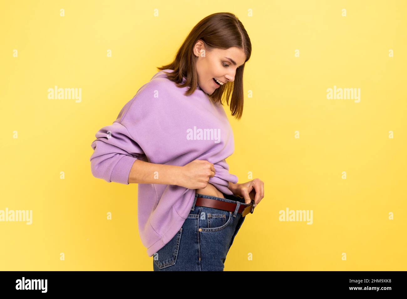 Seitenansicht der dunkelhaarigen glücklichen Frau, die erfolgreich abnimmt, Glück, Diät-Konzept zum Ausdruck bringt und einen violetten Hoodie trägt. Innenaufnahme des Studios isoliert auf gelbem Hintergrund. Stockfoto