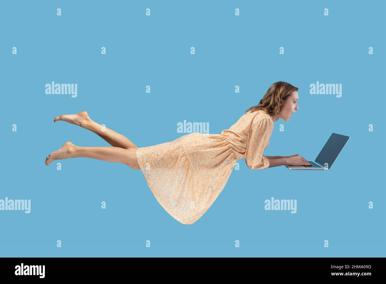 Schwebt in der Luft. Überrascht aufgeregt Mädchen gelben Kleid schweben mit Laptop, Tastatur eingeben, lesen schockierende Nachrichten auf Laptop-Bildschirm, während in der Luft fliegen. studio geschossen isoliert auf blauem Hintergrund Stockfoto