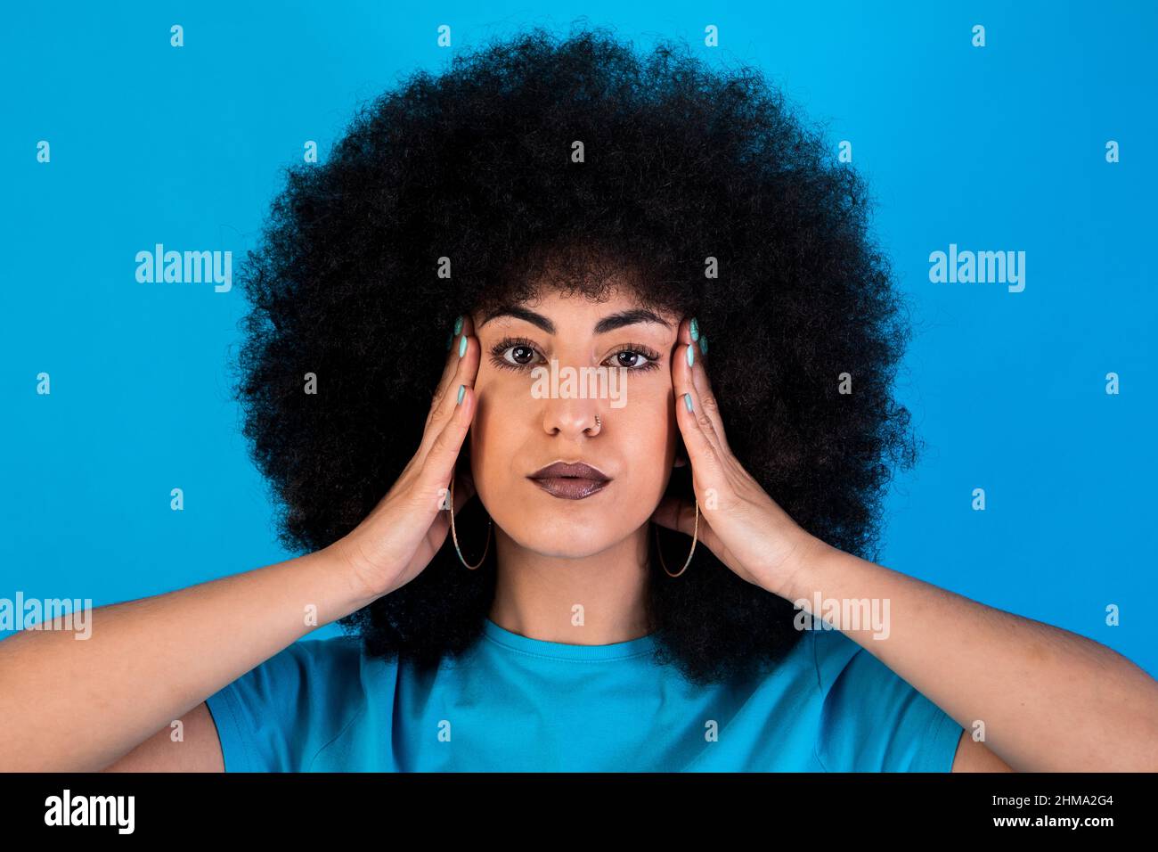 Seriöse hispanische Hündin mit schwarzer Afro-Frisur, die Tempel berührt und die Kamera anschaut, während sie auf blauem Hintergrund im Studio steht Stockfoto