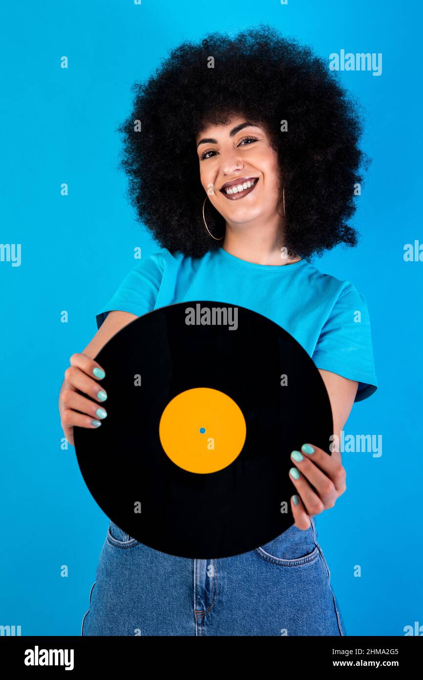 Lächelnd optimistische hispanische Meloman mit Retro-Vinyl-Schallplatte, die auf blauem Hintergrund die Kamera im Lichtstudio anschaut Stockfoto