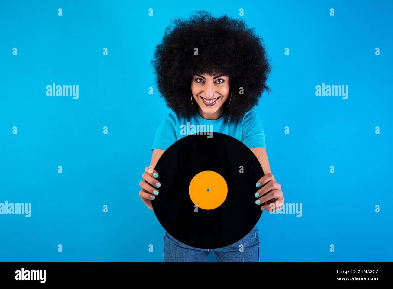 Lächelnd optimistische hispanische Meloman mit Retro-Vinyl-Schallplatte, die auf blauem Hintergrund die Kamera im Lichtstudio anschaut Stockfoto