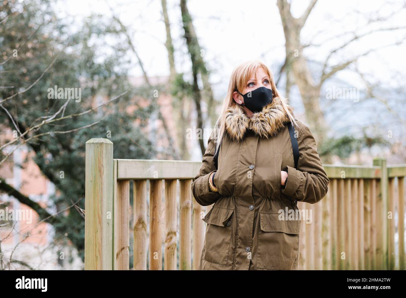 Frau in Oberbekleidung und schwarzer Schutzmaske, die während einer Pandemie am Herbsttag in der Stadt auf der Straße mit Bäumen und Holzzaun aufschaut Stockfoto
