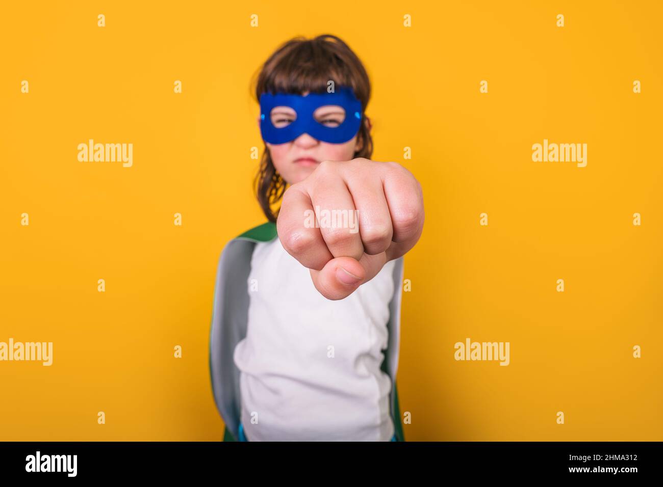 Selbstbewusstes Kind im Superhelden-Kostüm und Maske, die den Arm mit Faust vor der Kamera vor gelbem Hintergrund im Studio streckt Stockfoto