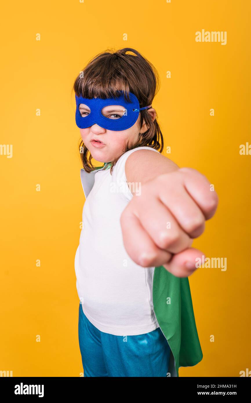 Selbstbewusstes Kind im Superhelden-Kostüm und Maske, die den Arm mit Faust vor der Kamera vor gelbem Hintergrund im Studio streckt Stockfoto