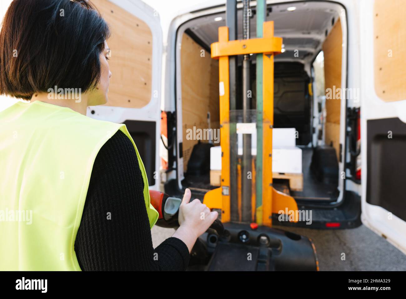 Seitenansicht einer nicht erkennbaren Lagerarbeiterin in grüner Uniform mit hohen Weste, die Ladeboxen in einen geöffneten LKW mit manuellem Stapler auf der Straße einlädt Stockfoto