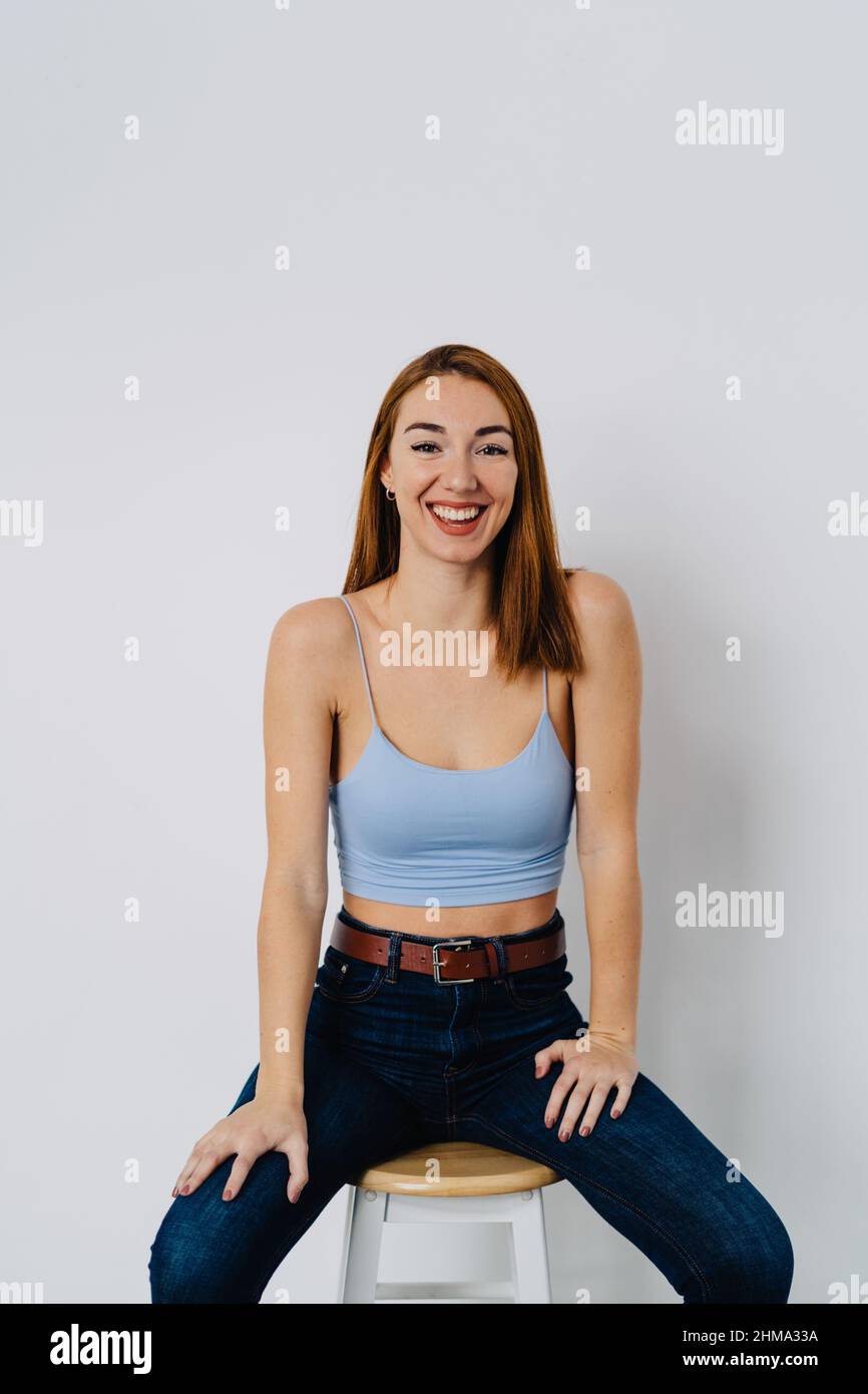 Positive junge Rothaarige, die Jeans und Oberteil trägt, sitzt auf einem Hocker nahe der weißen Wand und schaut auf die Kamera Stockfoto