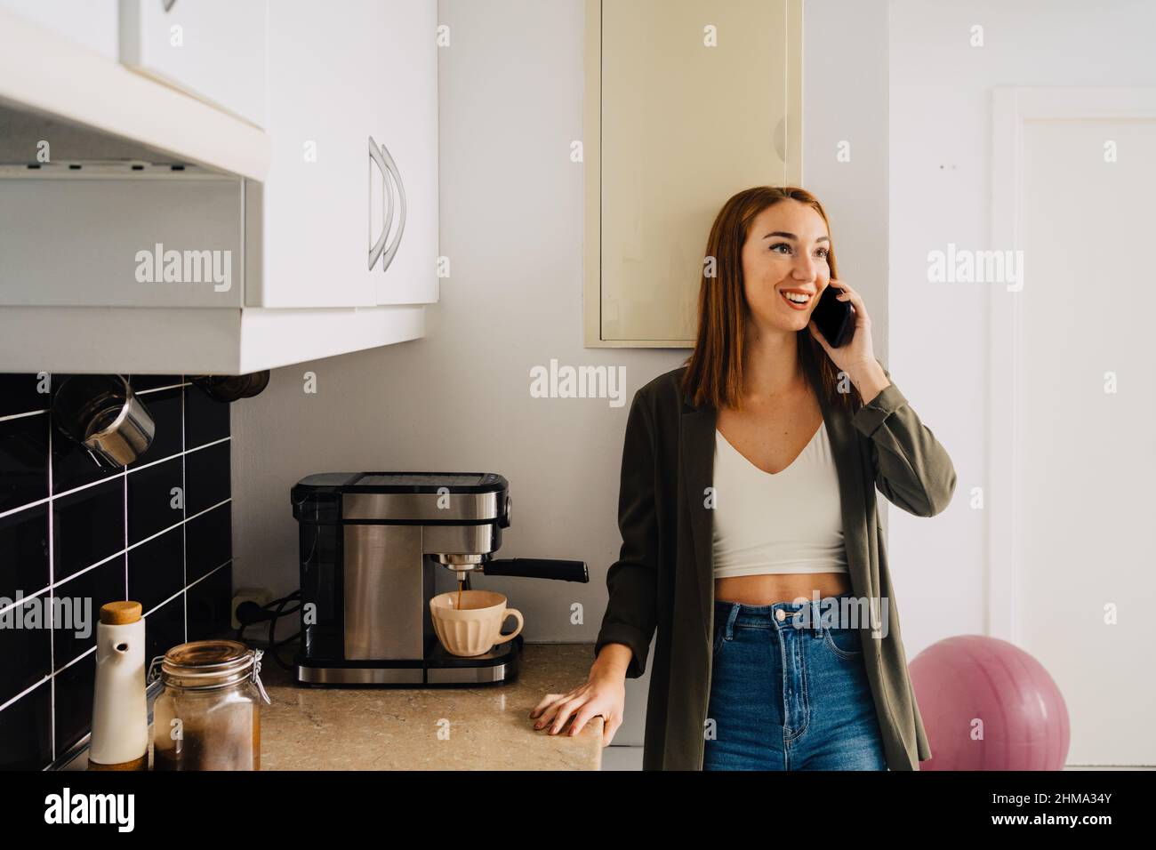 Positive Rothaarige Frau in legerer Kleidung, die auf dem Mobiltelefon spricht, während sie in der Küche mit einer Kaffeemaschine steht, die heißes Getränk in der Tasse zubereitet Stockfoto