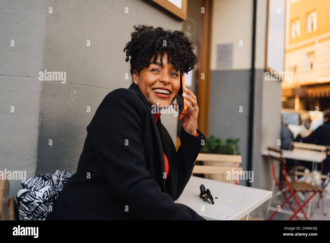 Aufgeregte, lockige afroamerikanische Frau in trendigem Outfit, die im Café im Freien am Tisch sitzt und Telefongespräche führt und wegschaut Stockfoto