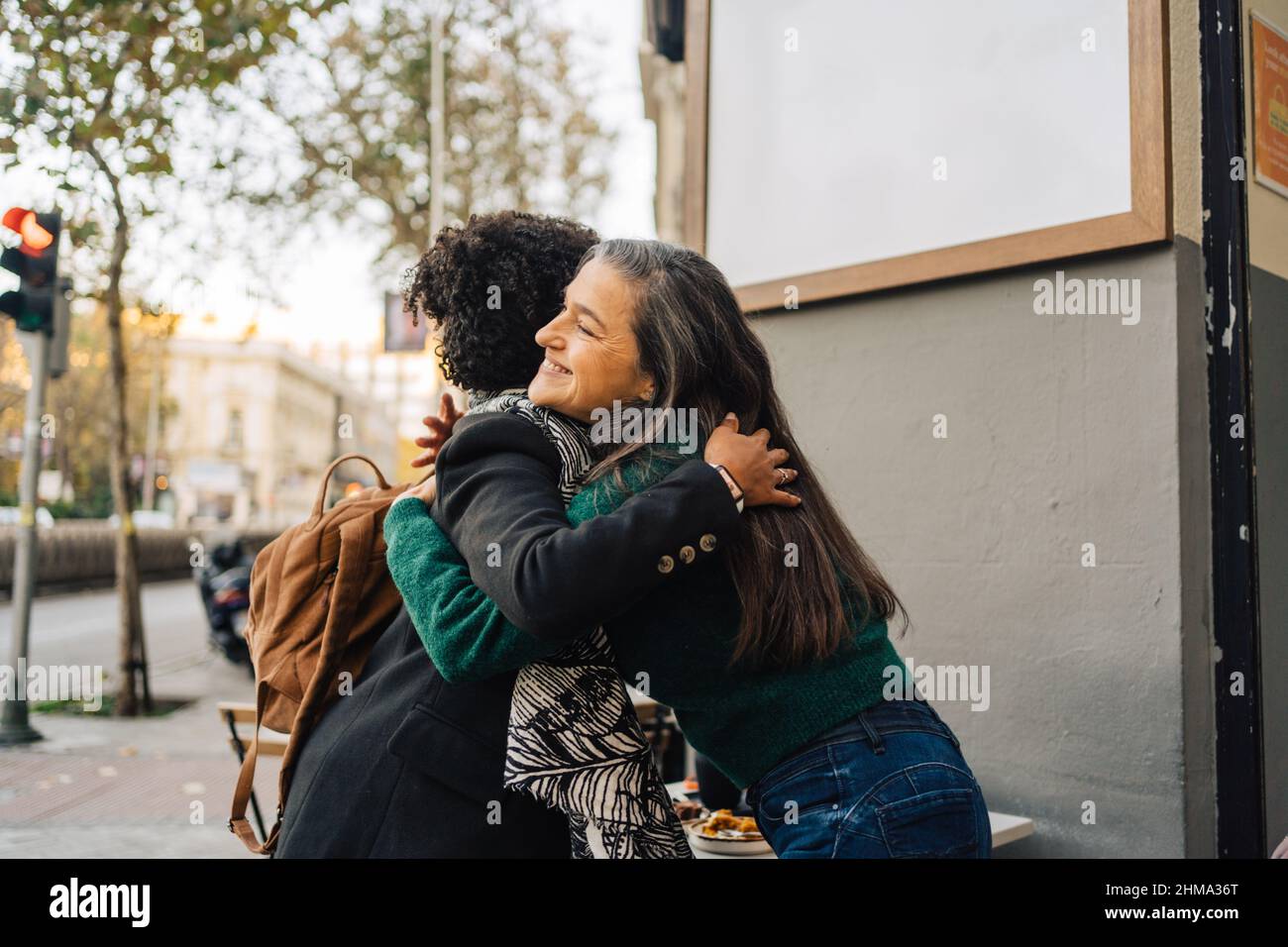 Seitenansicht einer lächelnden, reifen hispanischen Frau, die sich auf der Straße mit einer schwarzen, lockigen Freundin umarmt und sich gegenseitig begrüßt Stockfoto