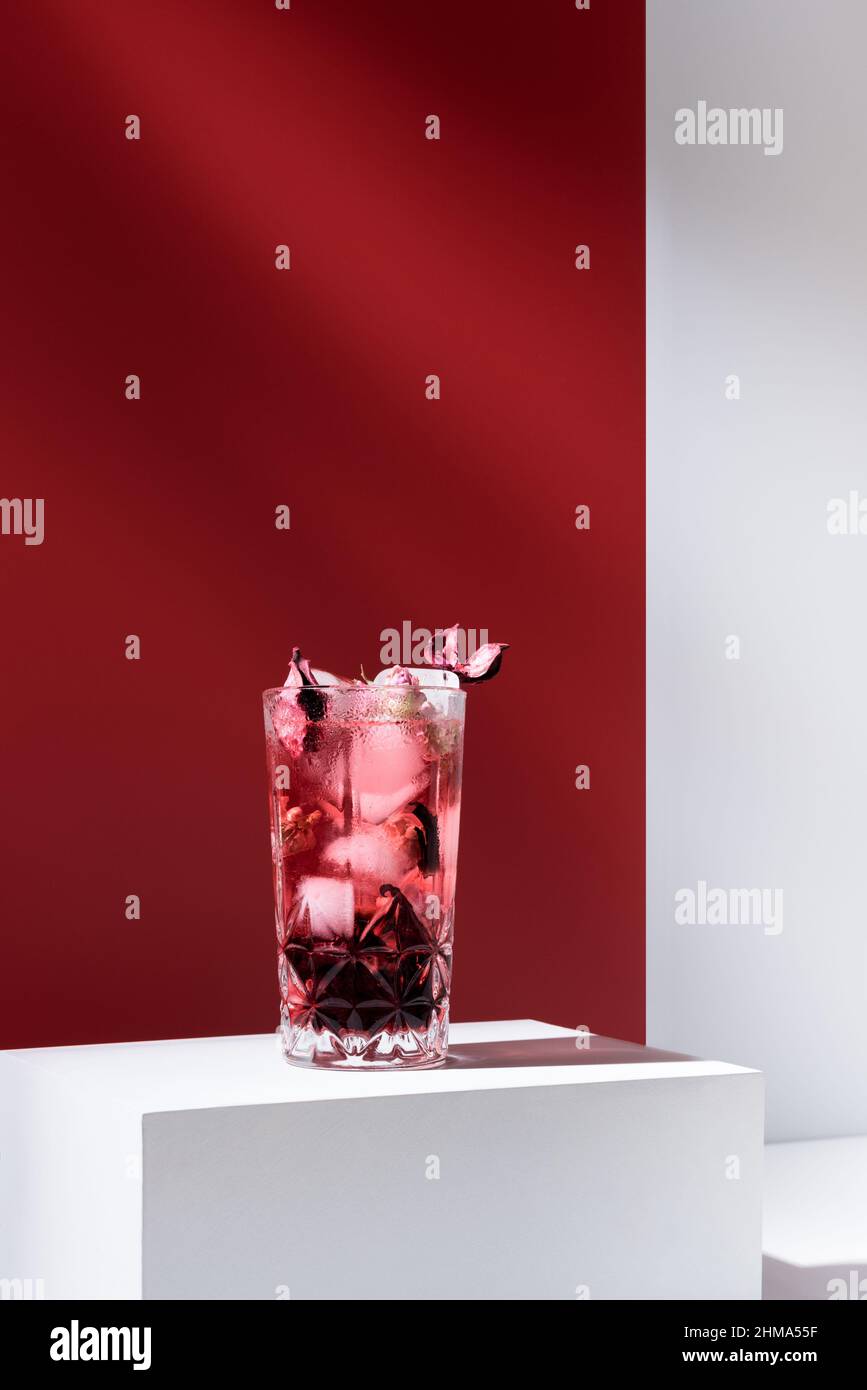 Komposition aus Glas gefüllt mit pinkem Gin Tonic und Eis auf weißem Tisch vor rotem Hintergrund Stockfoto