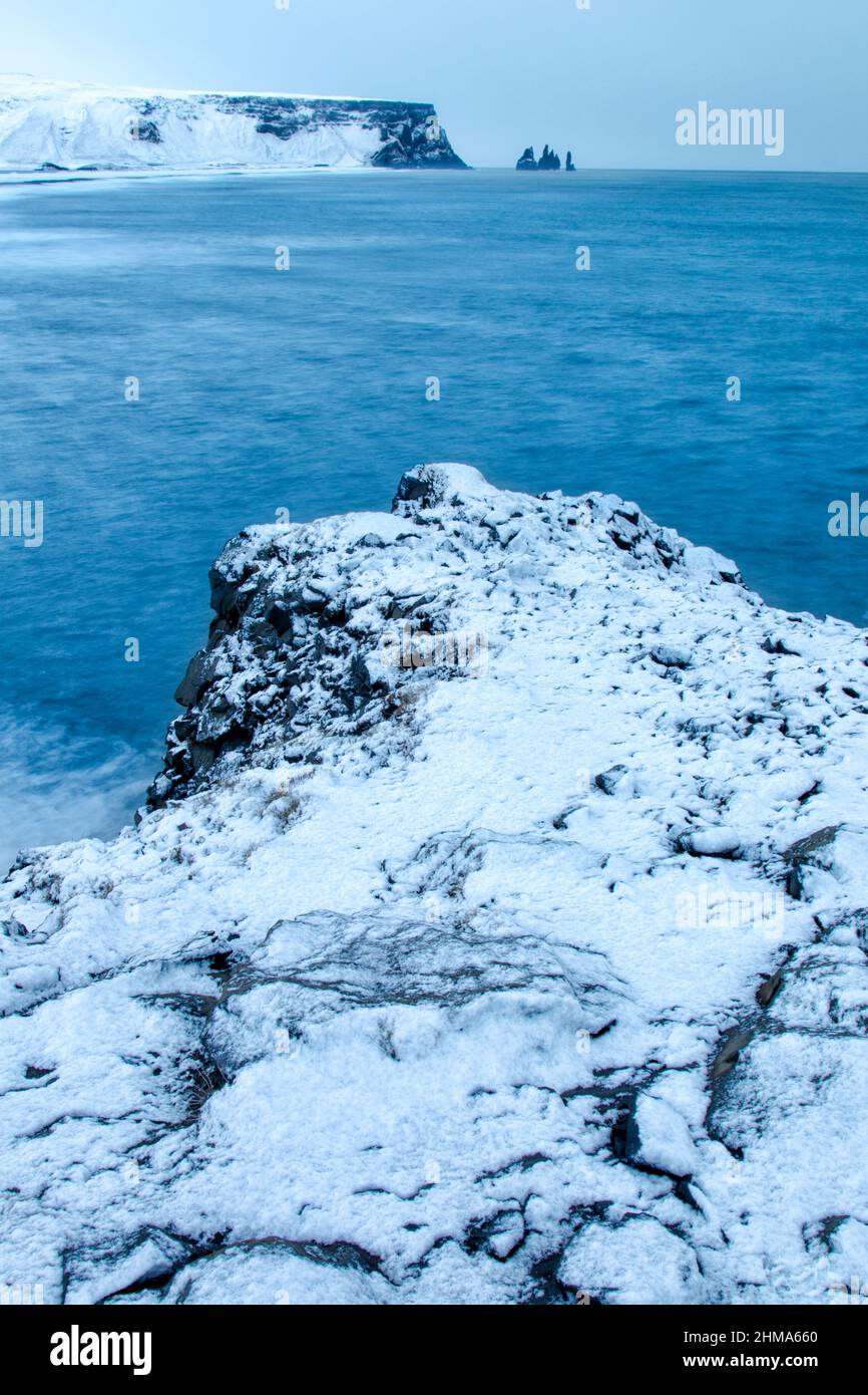 Schneebedeckte felsige Landzunge in der Nähe von Dyrhólaey mit Blick auf ferne Felsstapel bei Vik. Island. Stockfoto