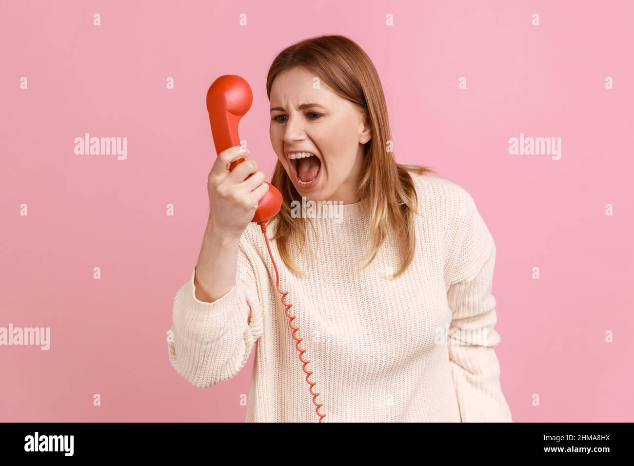 Portrait einer verärgerten blonden Frau, die in den roten Hörer schreit, ein unangenehmes Gespräch führt, Aggression ausdrückt und einen weißen Pullover trägt. Innenaufnahme des Studios isoliert auf rosa Hintergrund. Stockfoto