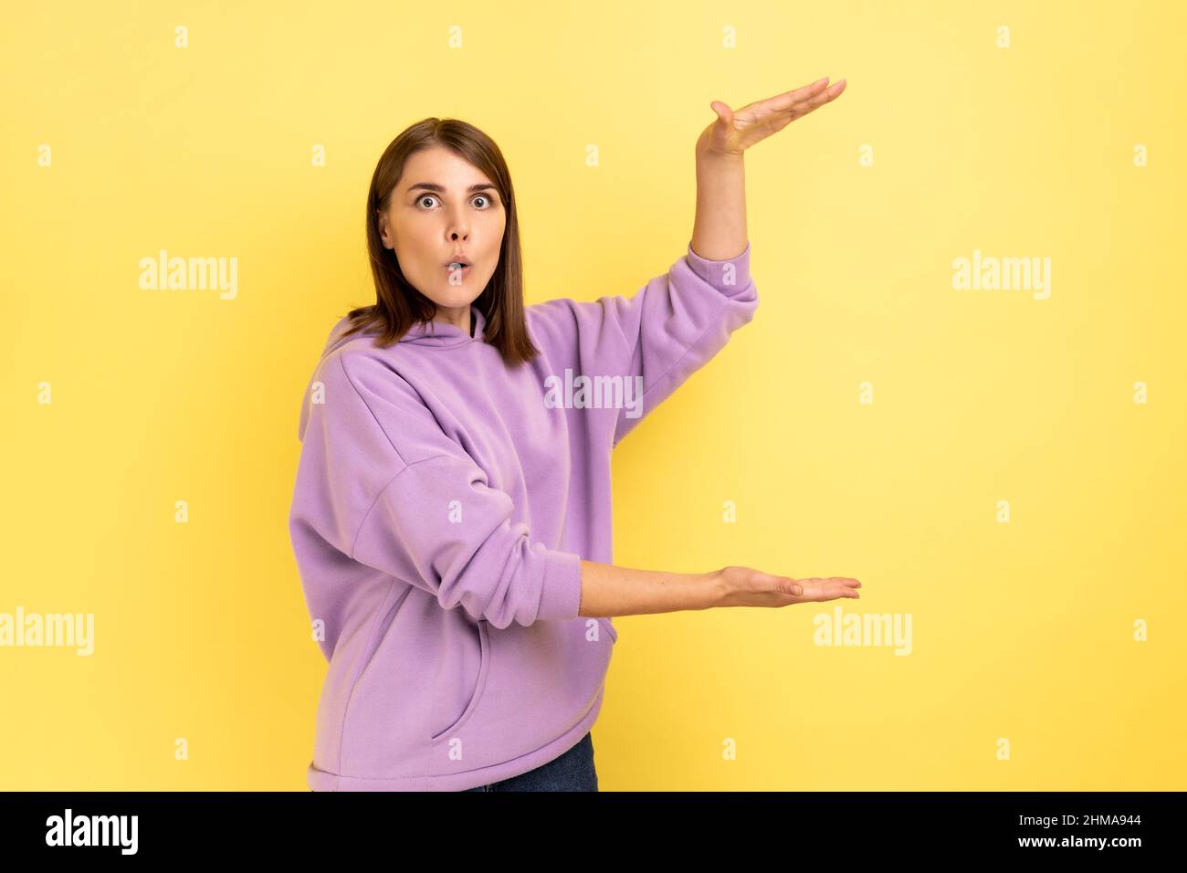 Porträt einer verblüfften, überraschten Frau, die einen Kopierraum zwischen den Händen präsentiert, einen leeren Platz für kommerzielle Texte hält und einen violetten Hoodie trägt. Innenaufnahme des Studios isoliert auf gelbem Hintergrund. Stockfoto