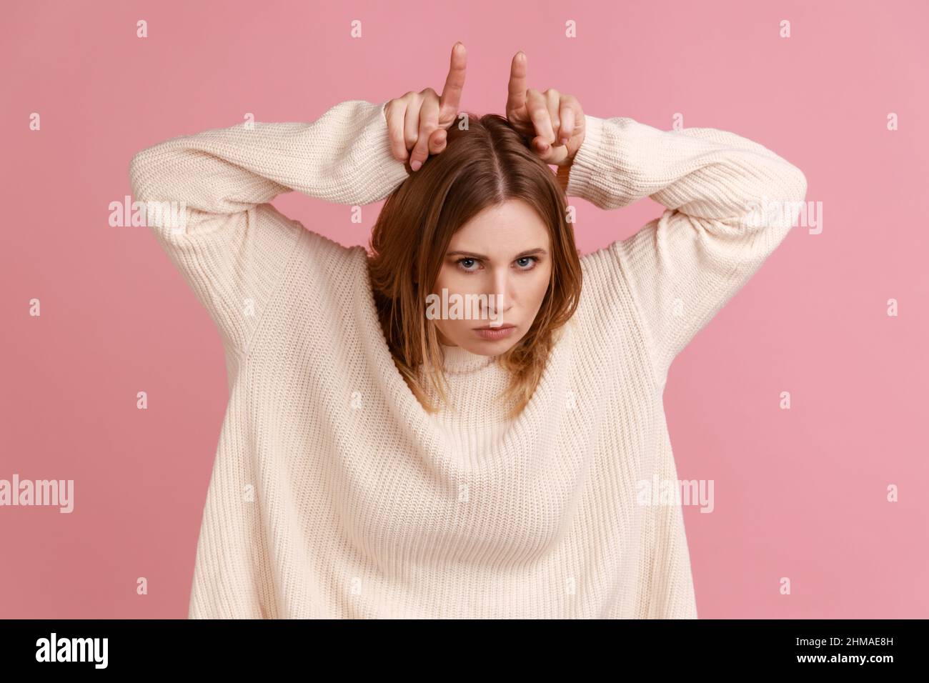 Porträt einer aggressiven blonden Frau, die Stierhörner über ihren Kopf und ihr Stirnrunzeln macht und die Bedrohung durch Wut zum Ausdruck bringt, in einem weißen Pullover. Innenaufnahme des Studios isoliert auf rosa Hintergrund. Stockfoto