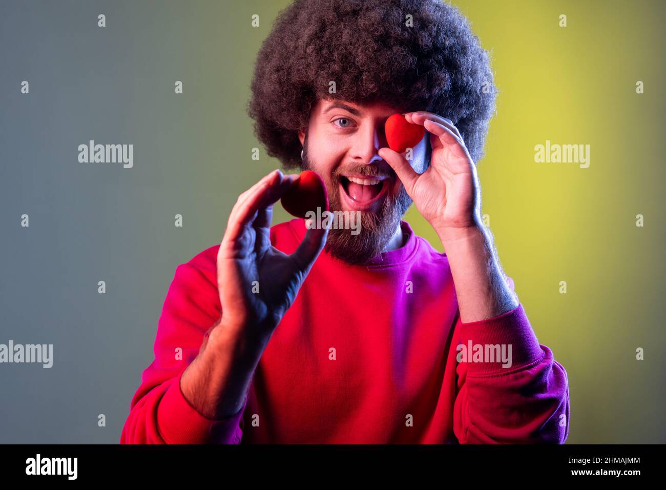 Hipster-Mann mit Afro-Frisur mit Spaß mit roten Spielzeugherzen, zeigt seine Zuneigung und Hingabe, Romantik, trägt rotes Sweatshirt. Innenaufnahmen im Studio, isoliert auf farbigem Neonlicht-Hintergrund. Stockfoto