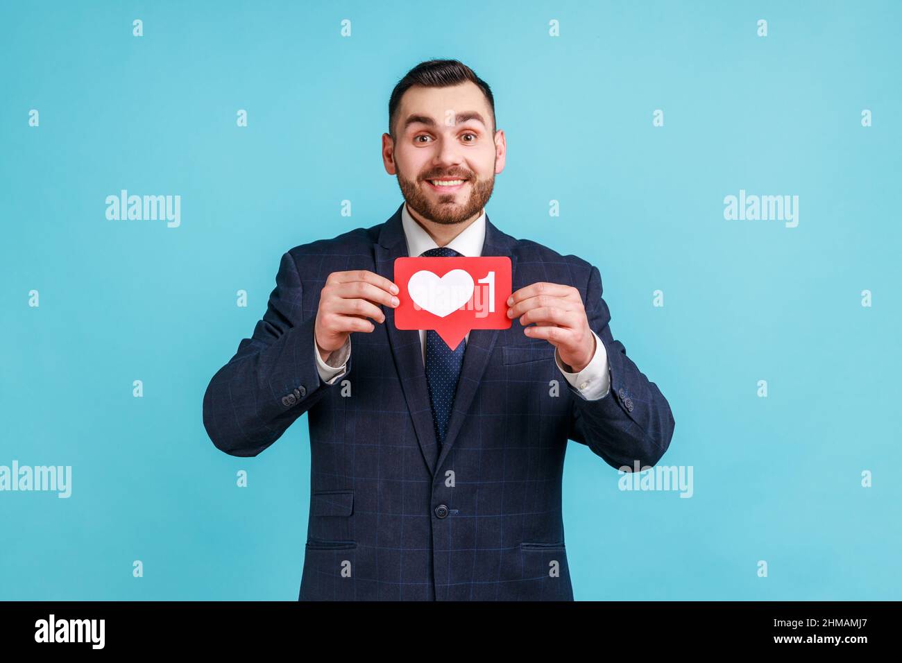Zufriedener Mann mit Bart trägt offiziellen Stil Anzug hält Herz-Symbol, empfehlen zu folgen und zu teilen Blog mit interessanten Inhalten. Innenaufnahme des Studios isoliert auf blauem Hintergrund. Stockfoto